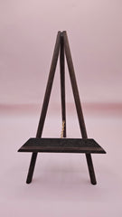 Rustic mini wood easel with dark shelf, set against a pink background. Vintage-style handmade display stand.