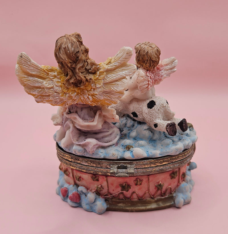 Whimsical vintage trinket box featuring a winged angel holding a basket of flowers, a cherub riding a spotted cow, and decorative star details on a round pastel base.