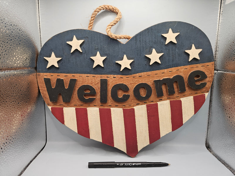 Close-up view of a 3D wood patriotic welcome heart door hanger, featuring an American flag design with raised stars and stripes. The door hanger measures 15"x10" and adds a bold, patriotic touch to any front door.