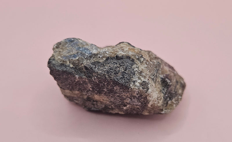 Close-up of a 106g Madagascar Labradorite Rough Specimen with natural gray matrix and subtle flashes of iridescent blue and green, displayed on a light pink background.