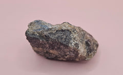 Close-up of a 106g Madagascar Labradorite Rough Specimen with natural gray matrix and subtle flashes of iridescent blue and green, displayed on a light pink background.