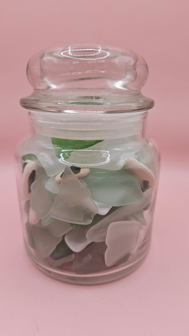 Apothecary glass jar filled with aqua and green sea glass and pottery shards, coastal decor accent on pink background