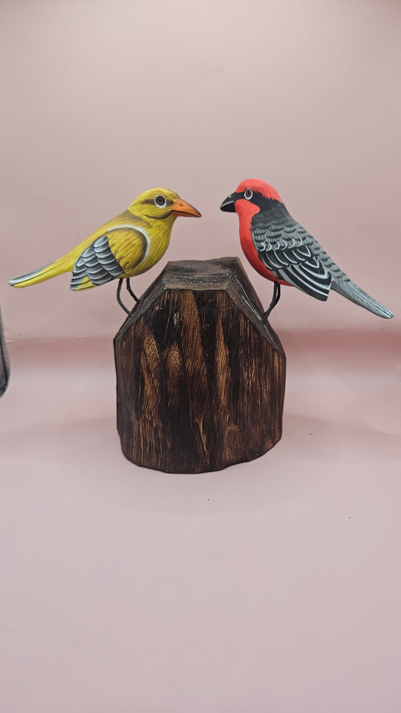 Hand-painted wooden songbird figurines in red/yellow and blue/yellow perched on rustic wood stumps, displayed on a green tabletop.