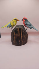 Hand-painted wooden songbird figurines in red/yellow and blue/yellow perched on rustic wood stumps, displayed on a green tabletop.