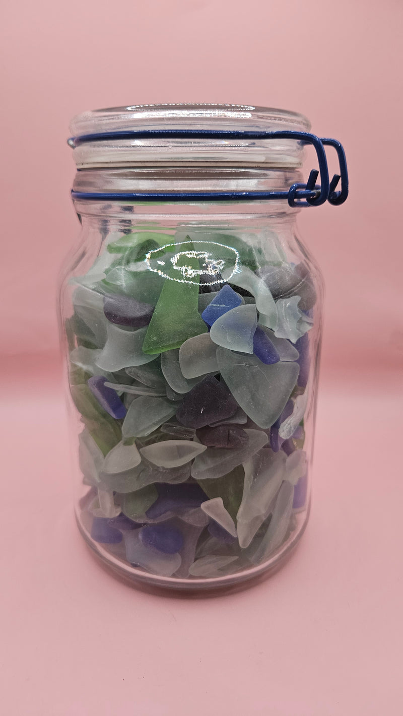 Clamp-lid glass jar with blue hinge filled with multicolored sea glass in green, blue, and frosted white, coastal home decor on pink background