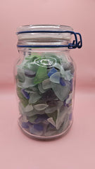 Clamp-lid glass jar with blue hinge filled with multicolored sea glass in green, blue, and frosted white, coastal home decor on pink background