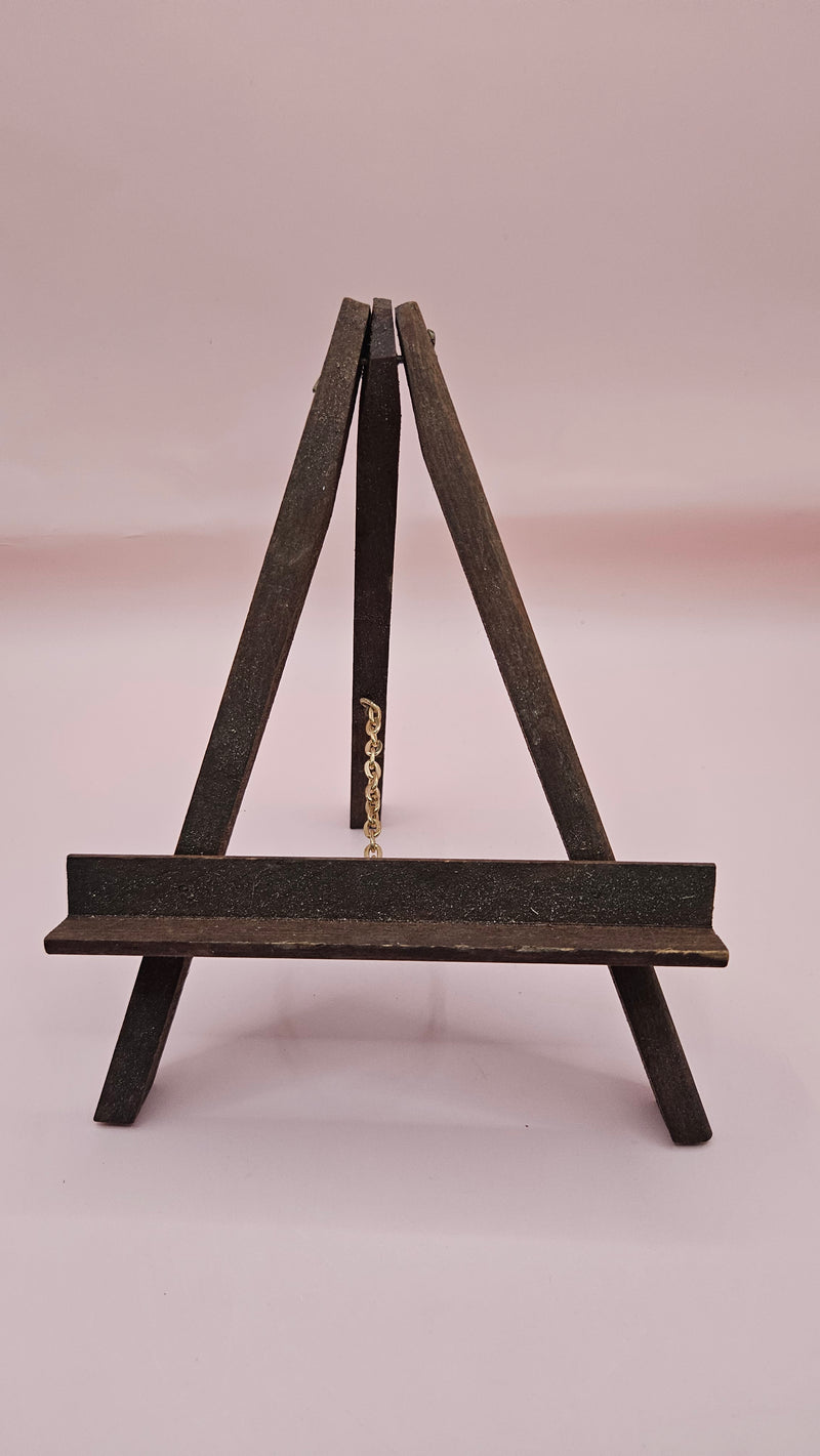 Rustic mini wood easel with dark shelf, set against a pink background. Vintage-style handmade display stand.