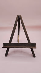 Rustic mini wood easel with dark shelf, set against a pink background. Vintage-style handmade display stand.
