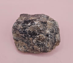 Close-up of a 106g Madagascar Labradorite Rough Specimen with natural gray matrix and subtle flashes of iridescent blue and green, displayed on a light pink background.