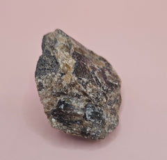 Close-up of a 106g Madagascar Labradorite Rough Specimen with natural gray matrix and subtle flashes of iridescent blue and green, displayed on a light pink background.