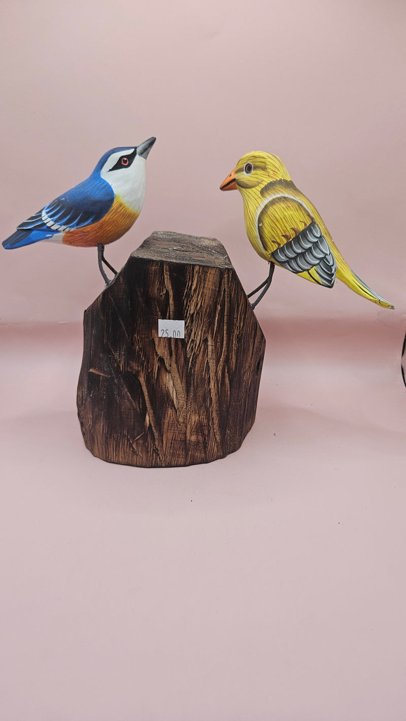 Hand-painted wooden songbird figurines in red/yellow and blue/yellow perched on rustic wood stumps, displayed on a green tabletop.