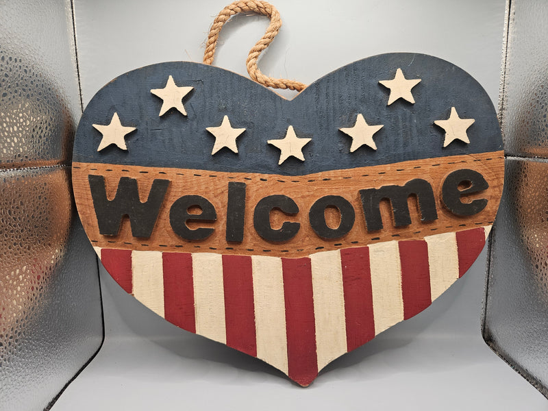 Close-up view of a 3D wood patriotic welcome heart door hanger, featuring an American flag design with raised stars and stripes. The door hanger measures 15"x10" and adds a bold, patriotic touch to any front door.