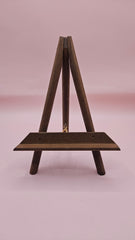 Rustic mini wood easel with dark shelf, set against a pink background. Vintage-style handmade display stand.