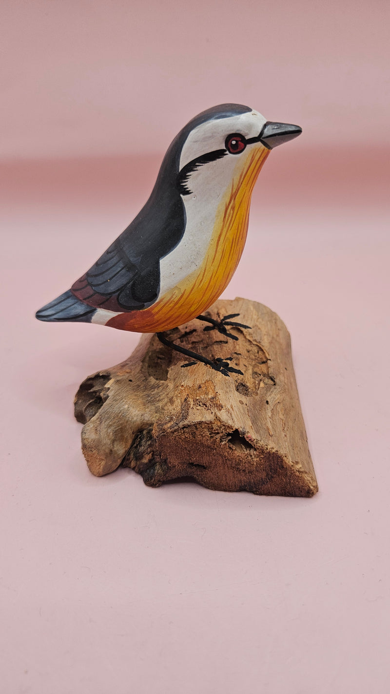 Photo of hand-carved wooden songbird figurine with grey, white, and yellow painted details, perched on a natural driftwood base.