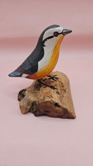 Photo of hand-carved wooden songbird figurine with grey, white, and yellow painted details, perched on a natural driftwood base.