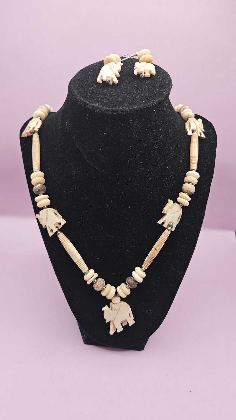 Vintage bone beaded elephant necklace (13") and earring set (1.5") featuring carved elephant pendants and earthy beads, displayed on a black stand with a soft pink background.