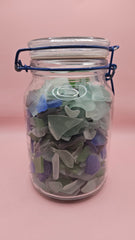 Clamp-lid glass jar with blue hinge filled with multicolored sea glass in green, blue, and frosted white, coastal home decor on pink background