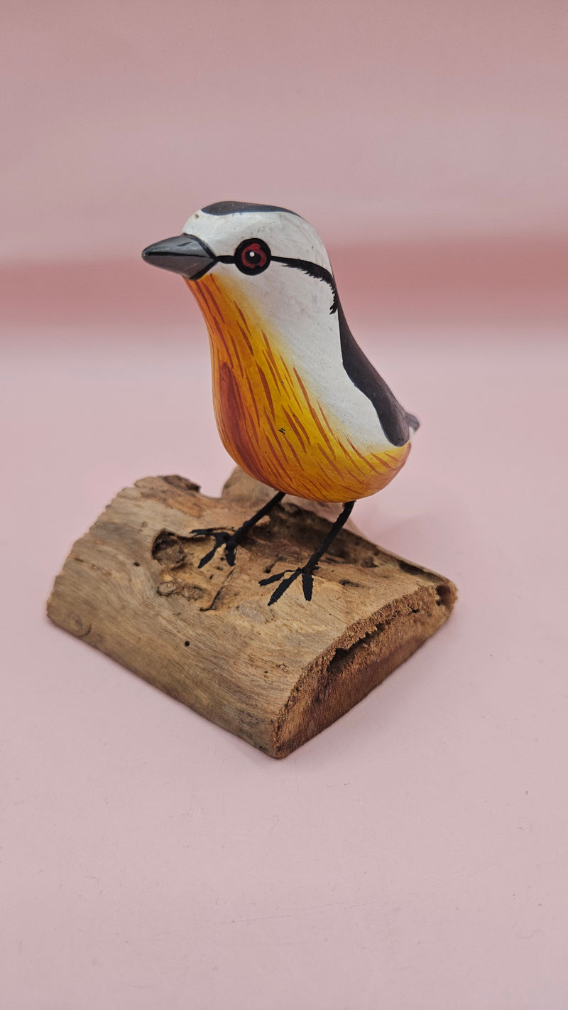 Photo of hand-carved wooden songbird figurine with grey, white, and yellow painted details, perched on a natural driftwood base.