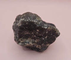 Close-up of a 270g Madagascar Labradorite Rough Specimen with natural gray matrix and subtle flashes of iridescent blue and green, displayed on a light pink background.