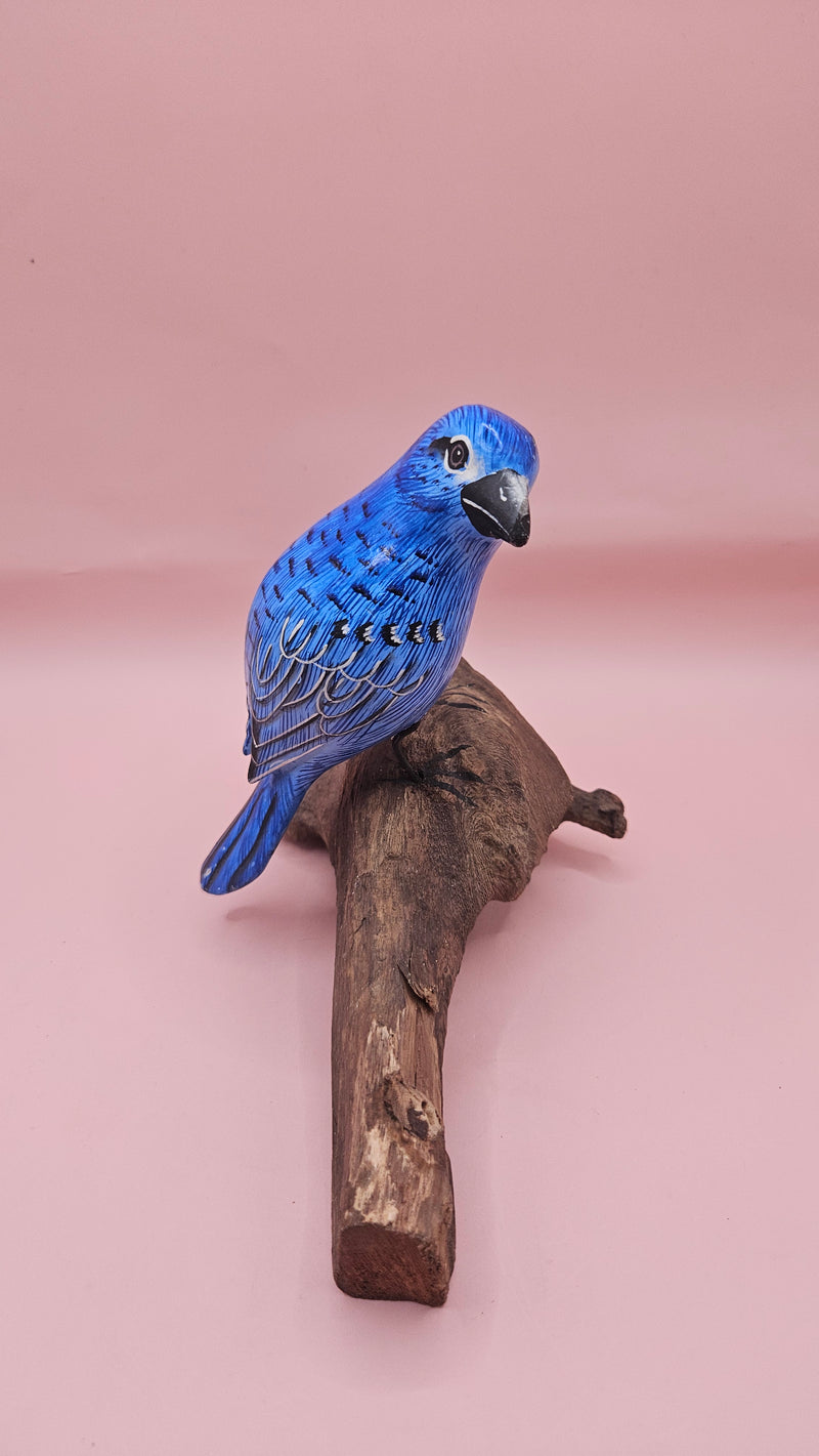 Hand-carved and painted blue bird sculpture perched on a natural driftwood base against a pink background.