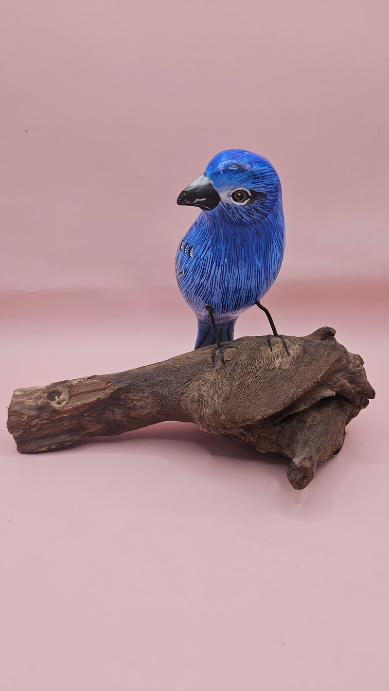 Hand-carved and painted blue bird sculpture perched on a natural driftwood base against a pink background.
