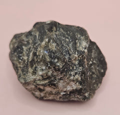 Close-up of a 270g Madagascar Labradorite Rough Specimen with natural gray matrix and subtle flashes of iridescent blue and green, displayed on a light pink background.