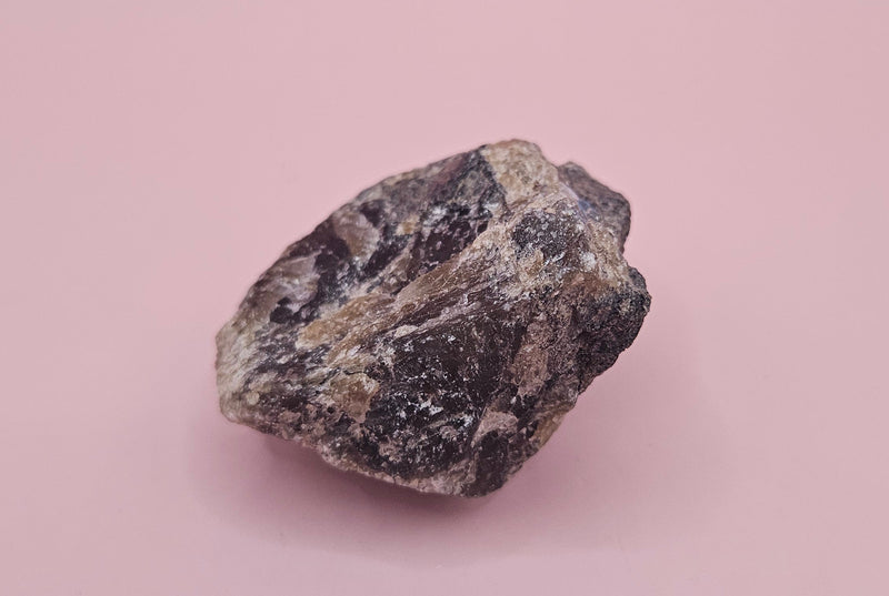 Close-up of a 106g Madagascar Labradorite Rough Specimen with natural gray matrix and subtle flashes of iridescent blue and green, displayed on a light pink background.