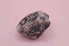 Close-up of a 106g Madagascar Labradorite Rough Specimen with natural gray matrix and subtle flashes of iridescent blue and green, displayed on a light pink background.