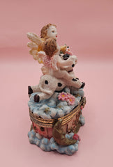 Whimsical vintage trinket box featuring a winged angel holding a basket of flowers, a cherub riding a spotted cow, and decorative star details on a round pastel base.
