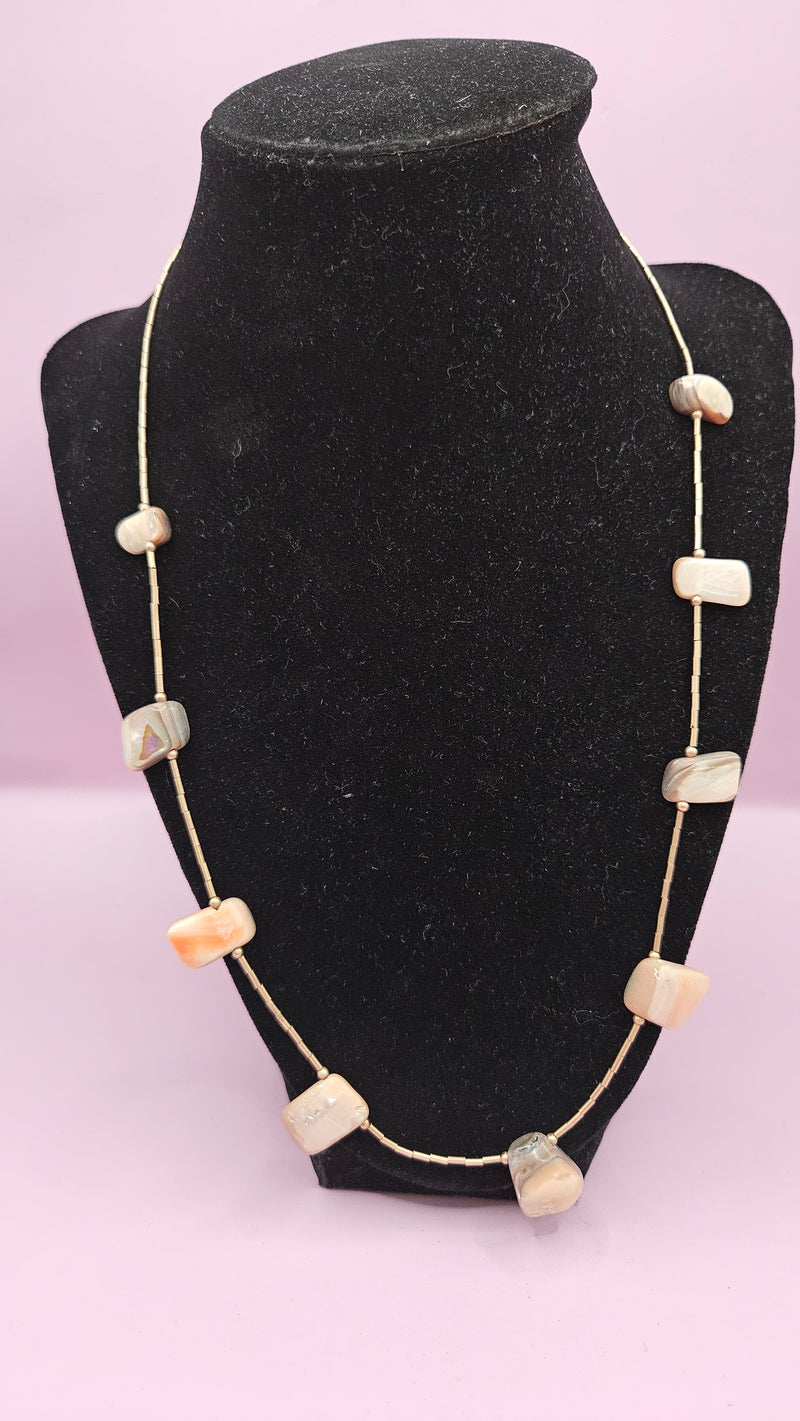 10.75" handmade natural shell necklace featuring iridescent and earthy shell beads on a delicate metallic chain, displayed on a black stand with a pink background.