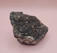 Close-up of a 270g Madagascar Labradorite Rough Specimen with natural gray matrix and subtle flashes of iridescent blue and green, displayed on a light pink background.