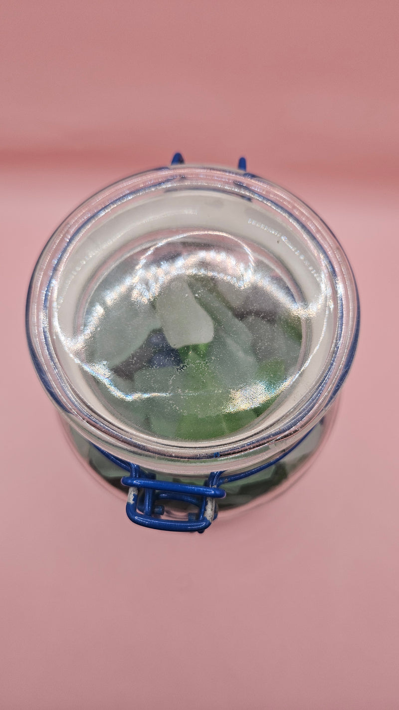 Clamp-lid glass jar with blue hinge filled with multicolored sea glass in green, blue, and frosted white, coastal home decor on pink background