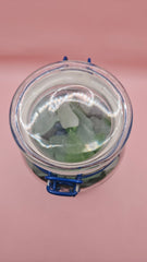 Clamp-lid glass jar with blue hinge filled with multicolored sea glass in green, blue, and frosted white, coastal home decor on pink background