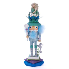 A HOLLYWOOD MERMAID KING NUTCRACKER featuring a seahorse and nautical-inspired design. The nutcracker is decorated with a blue and green mermaid sitting atop a green hat with a purple base, resembling a nautical chart.
