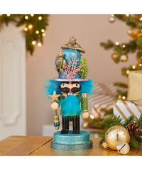 An 11-inch nutcracker figurine with a sea turtle hat, decorated with a blue-green color and accessories such as a star and a compass, placed on a table with Christmas decorations in the background.