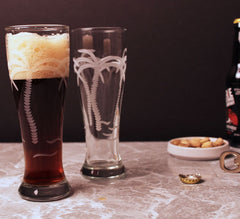 Two etched pilsner glasses featuring a palm tree design. One glass is filled with dark beer, while the other is empty, showcasing the intricate palm tree etching. Ideal for tropical or beach-themed settings.