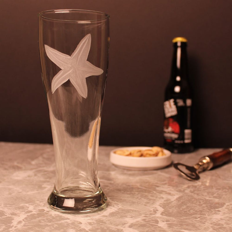 Etched pilsner glass featuring a large starfish design. The glass is set on a marble countertop with a bottle opener and snacks, perfect for a casual beer-drinking session.