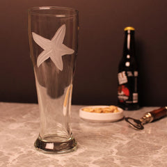 Etched pilsner glass featuring a large starfish design. The glass is set on a marble countertop with a bottle opener and snacks, perfect for a casual beer-drinking session.